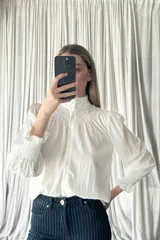 Camisa blanca de textura arrugada con cuello alto tipo gorguera, detalle de fruncido en el pecho y mangas largas abullonadas con puños elásticos fruncidos.