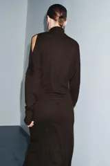 Vestido midi de punto color marrón, con cuello subido, manga larga y diseño cut out en un hombro. Presenta frunces laterales en la cintura que aportan un efecto drapeado.