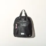 Mochila negra con doble asa de mano y tiras traseras regulables. Tiene un bolsillo frontal con cierre y un pequeño aplique rectangular en la parte inferior.
