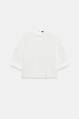 Camisa blanca de cuello redondo con mangas cortas abullonadas, detalles de frunces, puntillas y entredoses con cierre posterior.