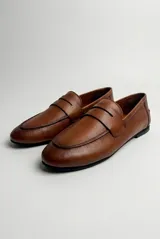Mocasín de cuero color café, diseño clásico de estilo penny loafer con suela plana y costuras visibles.