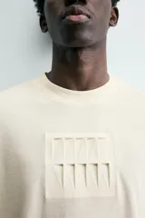 Camiseta de manga corta y cuello redondo, color crema o blanco roto, con un parche texturizado en el pecho que simula barras verticales.