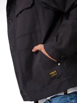 Chaqueta de lona color negro con forro interior de franela cepillada. Presenta cierre frontal con botones, puños abotonados, bolsillos en el pecho con solapa y bolsillos laterales. Incluye etiqueta de marca Caterpillar en el bolsillo inferior.