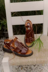 Sandalia de cuero vacuno color marrón, con diseño moderno de tiras anchas entrecruzadas, pasadores con cordón ajustable en el empeine y suela track robusta.