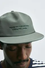 Gorra de cinco paneles color verde oliva con visera plana y ajuste trasero. Presenta un bordado frontal en negro con la frase "Time in art, music and coffee. Notting Hill, London".
