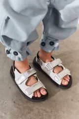 Sandalias de estilo deportivo con plataforma plana negra. Presentan un diseño de tiras anchas con cierre de velcro, combinando cuero texturizado blanco con detalles en cuero metalizado plateado.