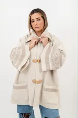 Saco tipo capa color beige, tejido en punto fino de lana con cierre de tancas de madera. Presenta cuello polo tejido y mangas anchas con detalles de trenzas y cintas tejidas.