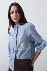 Camisa de rayas verticales azules y blancas con cuello mao y volados frontales. Presenta detalles de bordados florales y recortes con calados en el frente, con mangas largas abullonadas y cierre frontal con botones.