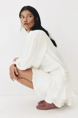 Vestido corto de color blanco en tejido con textura, con escote redondo, abertura con tiras para anudar, mangas largas abullonadas y falda de volados escalonados.