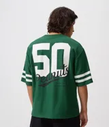 Remera de corte boxy y estilo deportivo, color verde oscuro, con diseño inspirado en el fútbol americano de Peanuts. Presenta cuello redondo, mangas cortas con franjas blancas horizontales y estampas localizadas de Snoopy en el frente.