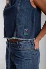Blusa sin mangas de denim lavado oscuro, con cuello redondo y costuras verticales contrastantes en color camel. Presenta un dobladillo deshilachado y un pequeño parche rectangular con diseño gráfico multicolor en la parte inferior frontal.