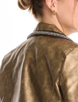 Chaqueta tipo biker de eco cuero con acabado metalizado dorado. Presenta cuello con solapa decorado con ribete bordado de pedrería y strass, cierre frontal asimétrico con cremallera metálica y bolsillos laterales funcionales con cierre.