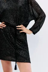 Vestido corto negro de manga larga con lentejuelas. Presenta escote redondo y un lazo ajustable en la cintura que frunce la falda. Las mangas son amplias y semitransparentes.