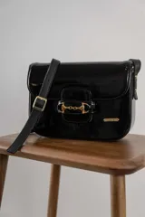 Cartera negra de cuero charolado con solapa y cierre de broche. Presenta un detalle metálico dorado en forma de freno de caballo en la parte frontal y una correa ajustable.