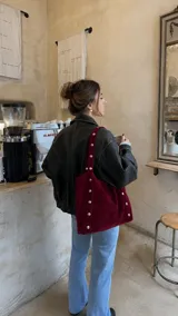 Cartera tipo tote bag color bordó, confeccionada en símil gamuza, con diseño amplio y detalle de tachas plateadas metálicas a lo largo de las tiras y los laterales.