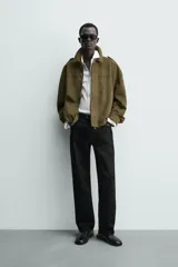 Cazadora cropped fit color verde militar, confeccionada en tejido efecto ante. Cuenta con cuello solapa, manga larga con puño abotonado, trabillas en los hombros, bolsillos laterales ocultos y un bolsillo interior. El bajo tiene elástico interior y el cierre frontal es con cremallera oculta por solapa con botones a presión.