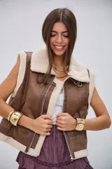 Chaleco de estilo aviador en color camel, confeccionado en material efecto cuero con interior y bordes de corderito sintético color crema. Presenta cierre frontal, cuello solapa y detalles de hebillas metálicas en los laterales y el cuello.