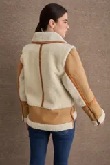 Campera tipo gamulán de cuero color camel con interior y detalles de piel sintética en color crema. Presenta cuello alto con doble hebilla ajustable, cierre frontal con cremallera y bolsillos laterales con cierre.
