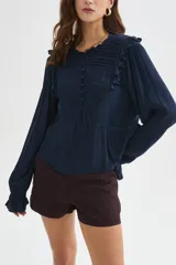 Blusa blanca de viscosa con cuello a la base, mangas largas abullonadas con puños ajustados, detalles de volados, encaje y botones frontales.