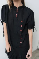 Vestido corto negro de broderie con cartera de botones asimétrica, mangas cortas abullonadas con lazo y volado en el ruedo.
