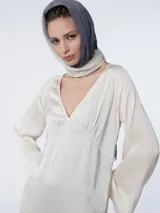 Vestido largo color crema, tipo túnica, confeccionado en crepé de satén de seda teñida en prenda. Tiene escote en pico con costura y ligeros fruncidos bajo el busto, líneas rectas y mangas acampanadas.