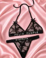 Conjunto de lencería de dos piezas confeccionado en encaje negro, compuesto por un soutiene triangular con tirantes halter y una bombacha tipo colaless. Presenta aplicaciones de letras brillantes con pedrería que forman la frase "FELIZ CUMPLE DISFRUTA".