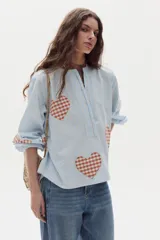 Camisa de algodón celeste a rayas verticales con parches estampados de corazones en tartán marrón y blanco. Posee cuello mao, cierre frontal parcial con botones y mangas largas abullonadas.