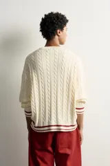 Jersey relaxed fit de punto tejido en hilatura de algodón, color crudo con detalles en bordó. Presenta cuello redondo, manga larga, diseño de trenzas y texto en intarsia a contraste en el delantero, con acabados en rib y líneas en los puños y el ruedo.