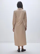 Vestido camisero largo color beige, con cuello clásico, mangas largas y cierre frontal con botones. Presenta un nudo decorativo a la altura de la cintura y una abertura en el ruedo.