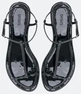 Sandalia baja negra de mujer con acabado en charol (verniz satinato). Diseño tipo thong (dedal) con tira fina que se une a una tira tobillera ajustable con hebilla metálica dorada.