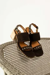 Sandalias de gamuza color marrón con taco cuadrado de madera. Presentan dos tiras anchas, una superior lisa y una inferior decorada con ojales metálicos plateados. Cuentan con cierre de pulsera con hebilla ajustable en el tobillo.