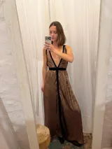 Vestido largo de seda color bronce con acabado brillante, escote profundo en V y espalda abierta. Presenta detalles en terciopelo negro que enmarcan el escote, recorren el frente y definen la cintura con una faja ajustable. Posee breteles anchos y una abertura frontal en la falda.