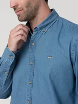 Camisa de manga larga color azul claro, confeccionada en tela tipo denim o chambray, con cuello abotonado (button-down) y un pequeño logo bordado de un pato en el pecho.