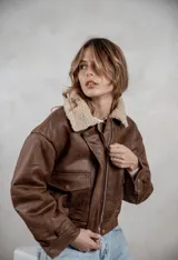 Campera de eco cuero color marrón con interior y cuello de corderito sintético. Presenta cierre frontal, bolsillos delanteros con solapa y diseño de corte bomber.