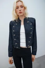 Chaqueta de denim de corte corto, color azul oscuro, con cuello redondo, hombreras y cierre frontal con cremallera. Presenta diseño de inspiración militar con doble botonadura metálica decorativa en el frente, trabillas con botón en los hombros y bajo elástico.