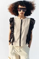 Blusa de cuello redondo y manga sisa, de color beige con rayas verticales. Presenta volantes negros en las sisas y una tira de encaje central con jaretas. Cuenta con cierre en la espalda con abertura y botón.