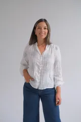 Blusa blanca de manga larga con diseño de rayas verticales y bordados florales en tono grisáceo. Presenta cuello camisero, cierre frontal con botones y puños elásticos.