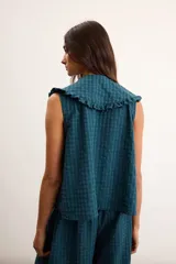Blusa de algodón con patrón a cuadros vichy en tonos verde oscuro y azul, sin mangas, con cuello amplio decorado con volados, botones delanteros y un bolsillo frontal aplicado con ribete de volados.
