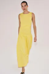 Vestido largo amarillo de algodón, con cuello barco y mangas sisa. Presenta diseño drapeado en los laterales y falda de corte evasé con volumen en el bajo.