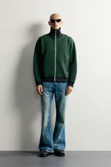 Chaqueta regular fit color verde oscuro con detalles en negro en el cuello subido, los puños, el dobladillo inferior y los ribetes de los bolsillos. Cuenta con cierre frontal de cremallera metálica y bolsillos con cierre en la cadera.