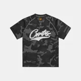 Camiseta de entrenamiento Corteiz con diseño de camuflaje en tonos grises y negros. Presenta cuello en V, mangas cortas y el logo de la marca estampado en el frente en color blanco.