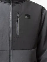 Campera de polar negro con cuello alto y cierre frontal completo. Presenta paneles de nylon en contraste en el pecho, hombros y codos, incluyendo un bolsillo vertical con cierre en el pecho y dos bolsillos laterales abiertos.