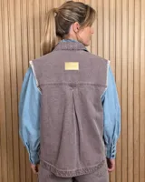 Chaleco de denim color marrón, confeccionado en 100% algodón, con bolsillos frontales amplios, cierre de botones y costuras decorativas bordadas en blanco en los bordes y los bolsillos.
