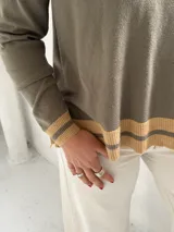 Sweater de punto en color verde oliva, con cuello en V y diseño holgado. Presenta detalles de franjas en color beige en el cuello, los puños y el ruedo.