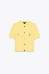 Chaqueta de punto color amarillo pálido, con cuello redondo, manga corta, cierre frontal con botones dorados y bolsillos de parche en el pecho.