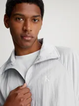 Campera deportiva Calvin Klein color gris claro, de sarga de satén brillante, con cuello alto, mangas raglán, puños y bajo elásticos, cierre de cremallera, bolsillo con cremallera en el pecho, bolsillos con botón de presión en la cintura, forro de malla transpirable y logo monograma estampado en el pecho.