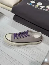 Zapatillas bajas de lona color gris con cordones violetas, puntera de goma blanca y suela de goma color crema con detalles negros.