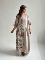 Vestido largo beige de lino con diseño midi holgado, cuello redondo, mangas cortas con vuelta y un gran estampado lateral de flores y hojas en tonos marrones y verdes. Cuenta con un bolsillo frontal aplicado con detalles deshilachados.