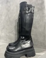 Bota bucanera negra de cuero sintético, con caña alta, plataforma y suela track. Presenta dos correas con hebillas plateadas en la parte superior de la caña.