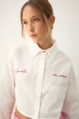 Camisa blanca de popelina de algodón, de corte cropped, con cuello clásico y cierre frontal de botones. Presenta bordados rojos en el frente con las frases "el poder" y "DEL AMOR", y un bolsillo de parche en el lado derecho.